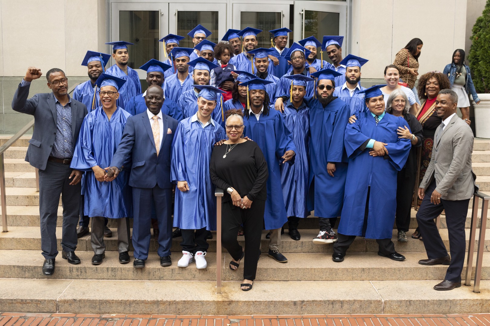 A group of graduates in blue hats and gowns cheer on the steps of Uris Hall