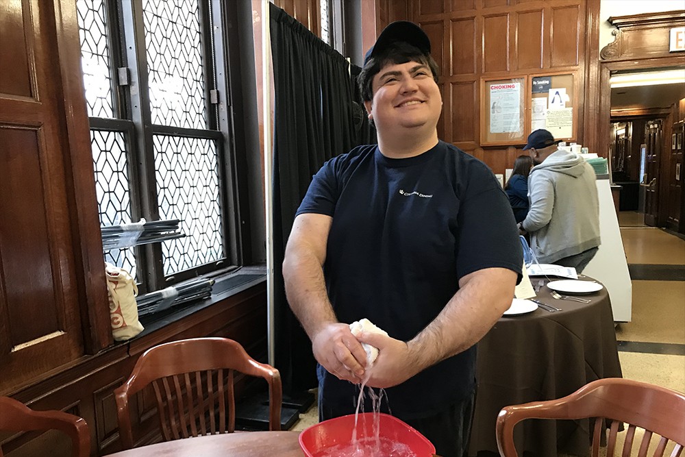 A SNACK employee smiles at a dining table he is about to wash with a rag.