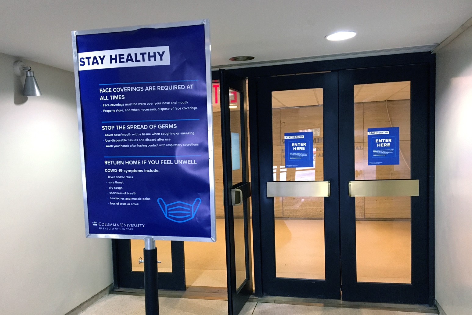 A large blue sign that reads "stay healthy" in front of the IAB doors that have smaller blue signs posted on the glass that say enter here