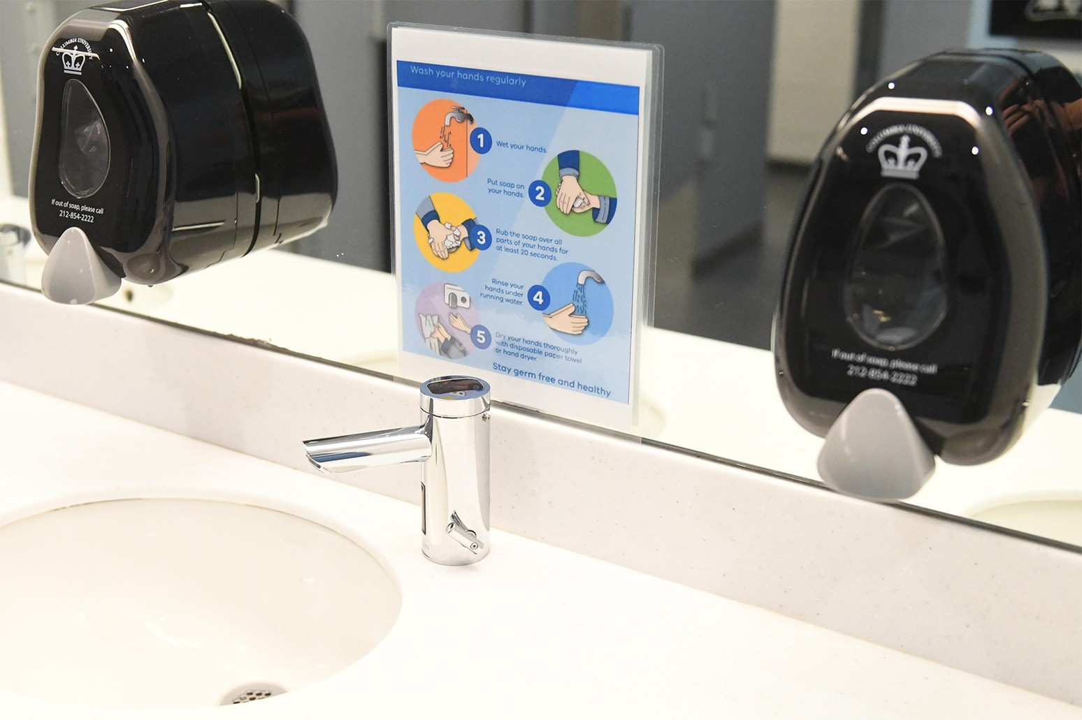 A restroom sink with a touchless faucet, two black soap dispensers, and a sign stating how to wash hands