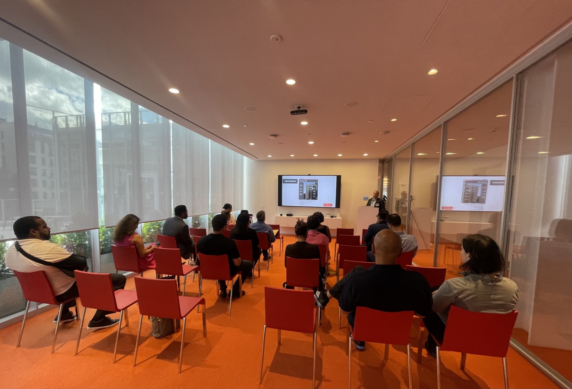 A room with tall windows, 15 participants sitting in red chairs and a presenter standing at the front of the room in front of a large tv screen with powerpoint presentation.