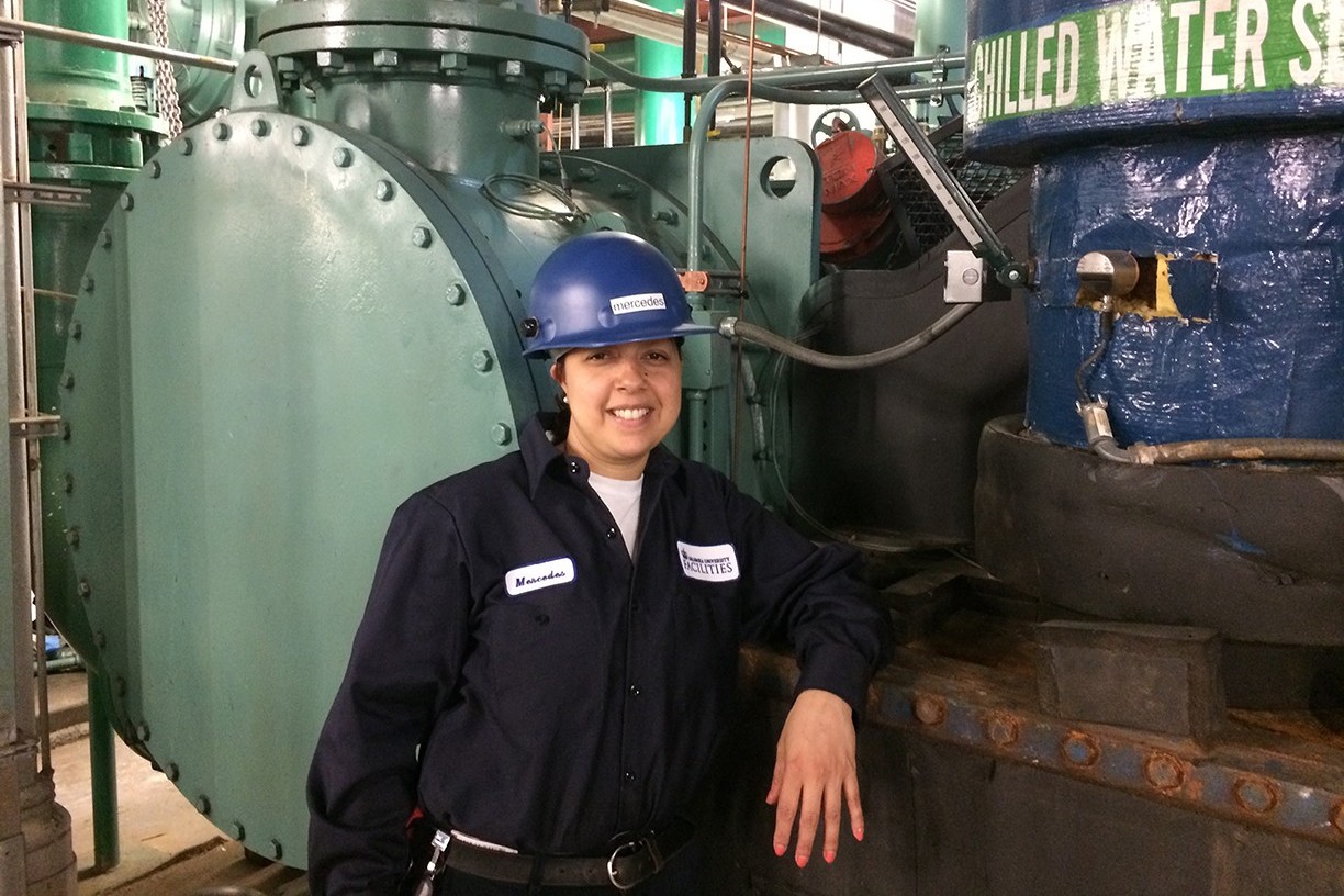 A woman with a hard hat stands next to chillers in a power plant.
