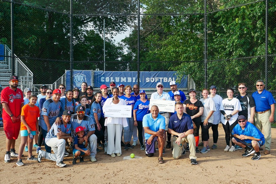 A group of people stand together at a baseball diamond holding large checks for charity.