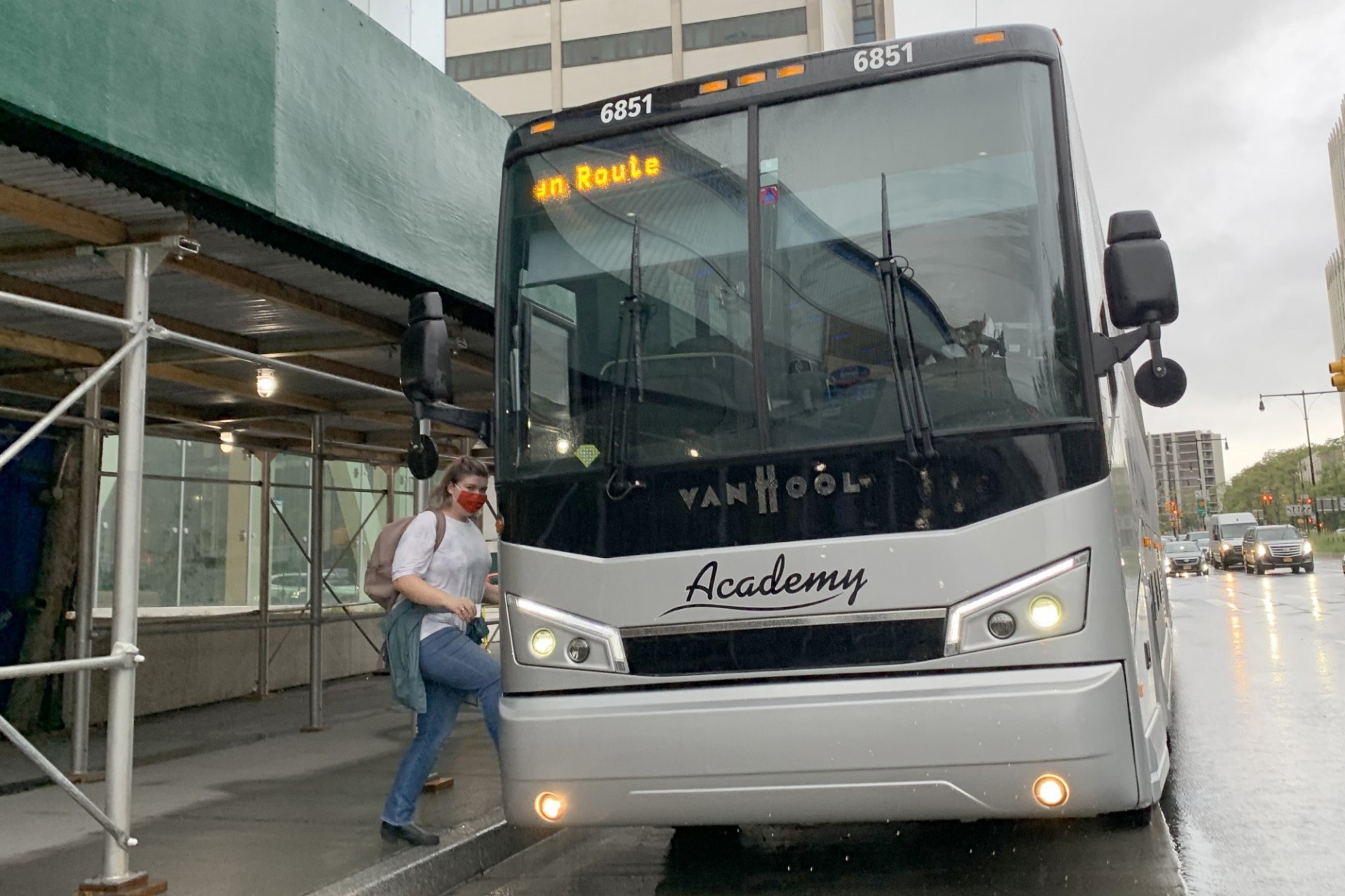 A person gets on a large gray Academy bus that has "CU Brooklyn" written on it from a sidewalk with sidewalk shedding.