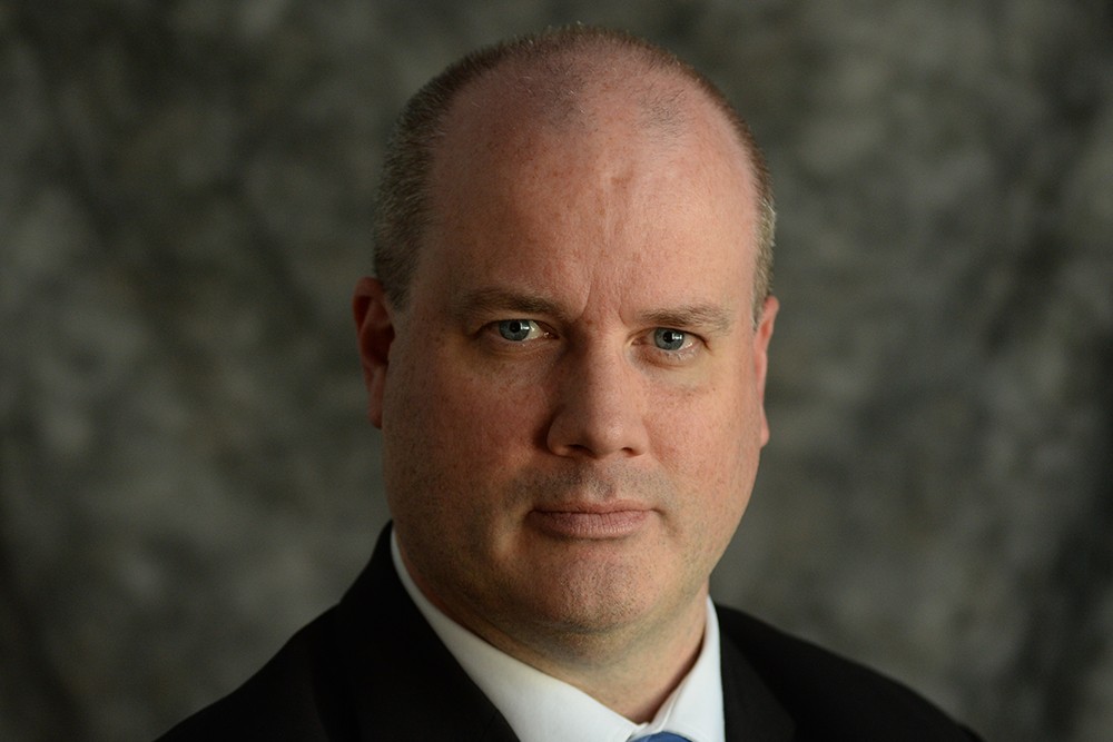 A headshot of Edward McArthur who is wearing a black suit and blue tie, against a gray background.