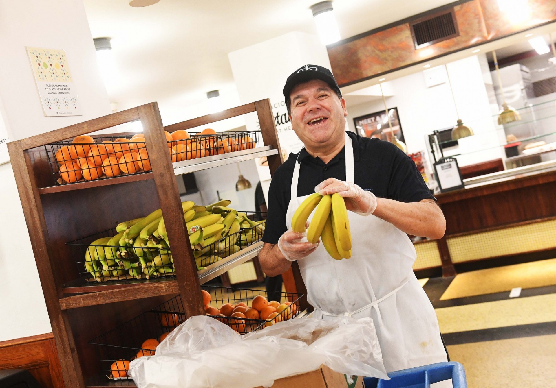 A man in a Columbia-branded black baseball cap, black polo shirt, and white apron holds a bunch of bananas in gloved hands.