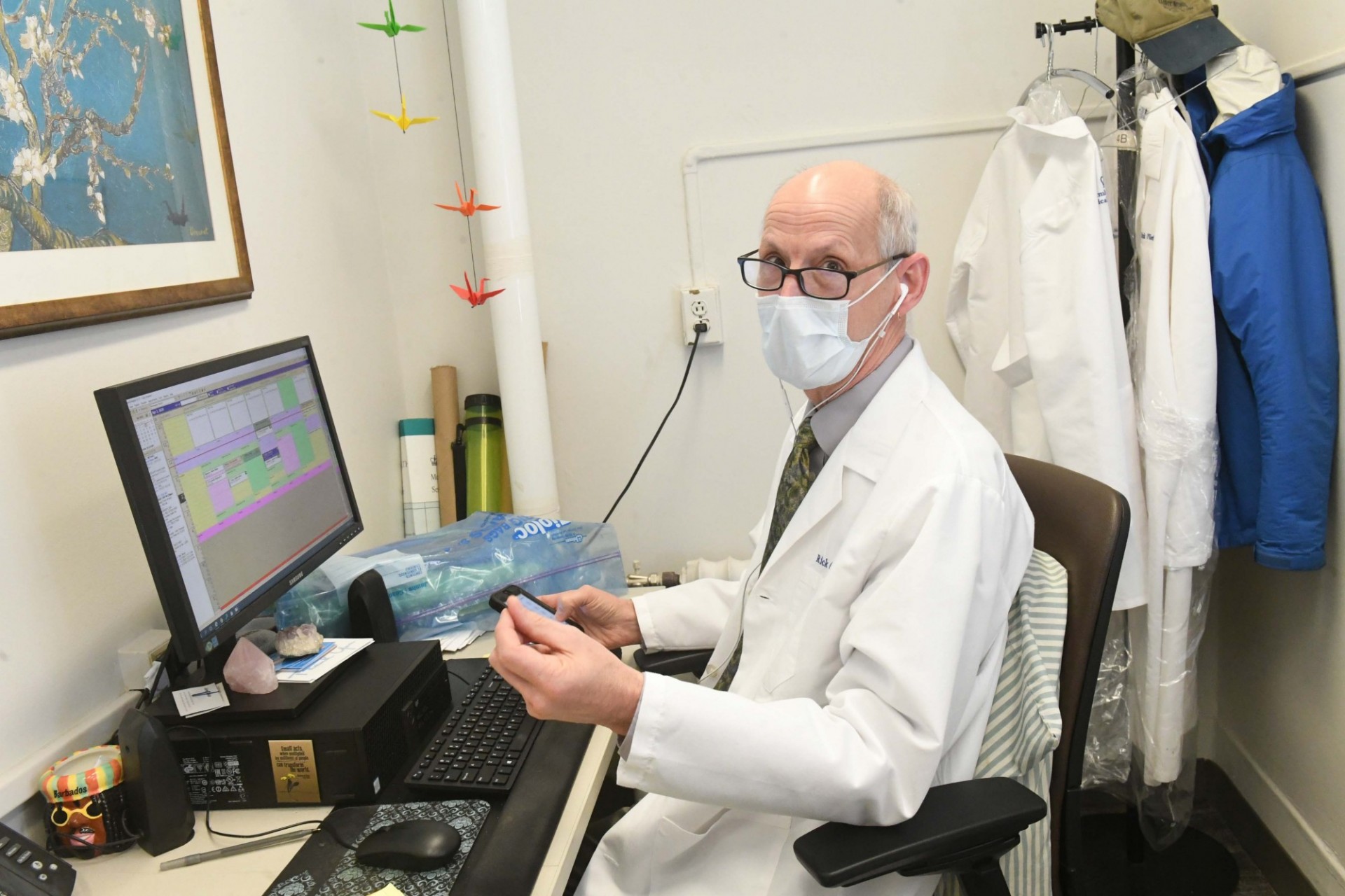 A man with black frame glasses wearing a mask and white coat sits in front of a computer while holding a smart phone.