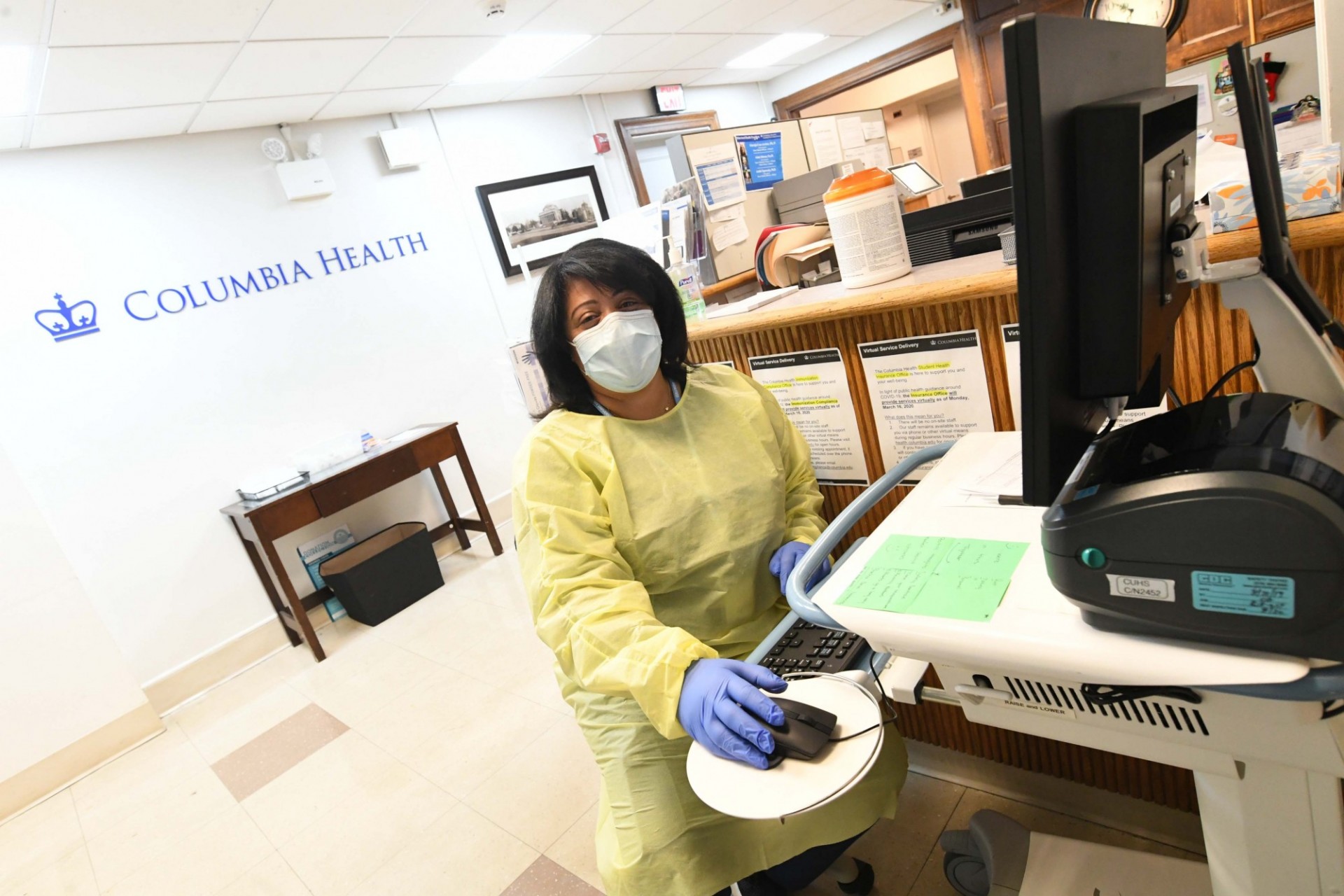 A member of the Health team is prepared to check-in students as they arrive to Medical Services. Photo credit Eileen Barroso. A member of the Health team wearing PPE sits at a computer monitor.