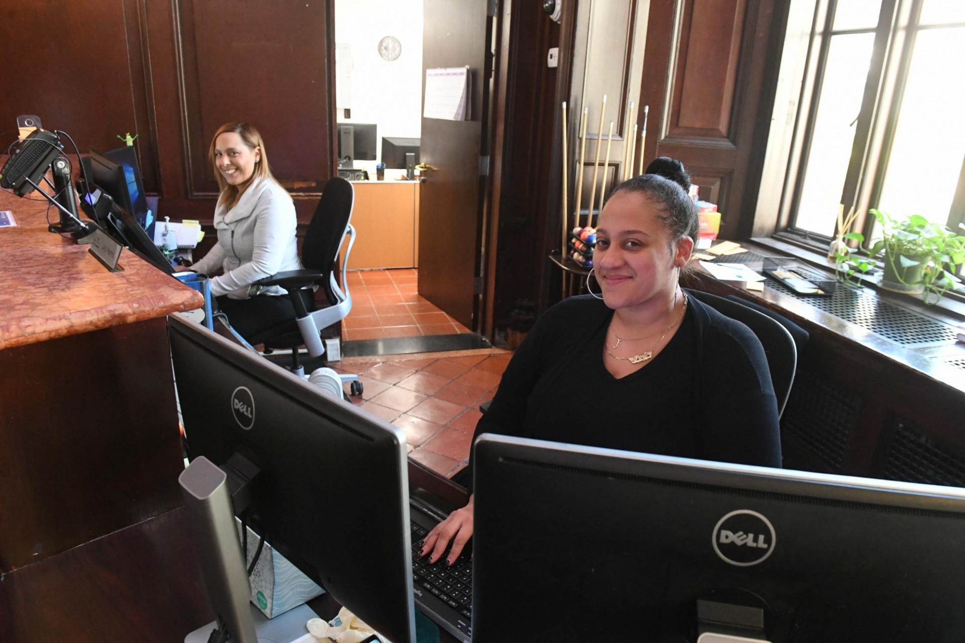 Two women sit in front of computer monitors behind a raised desk.