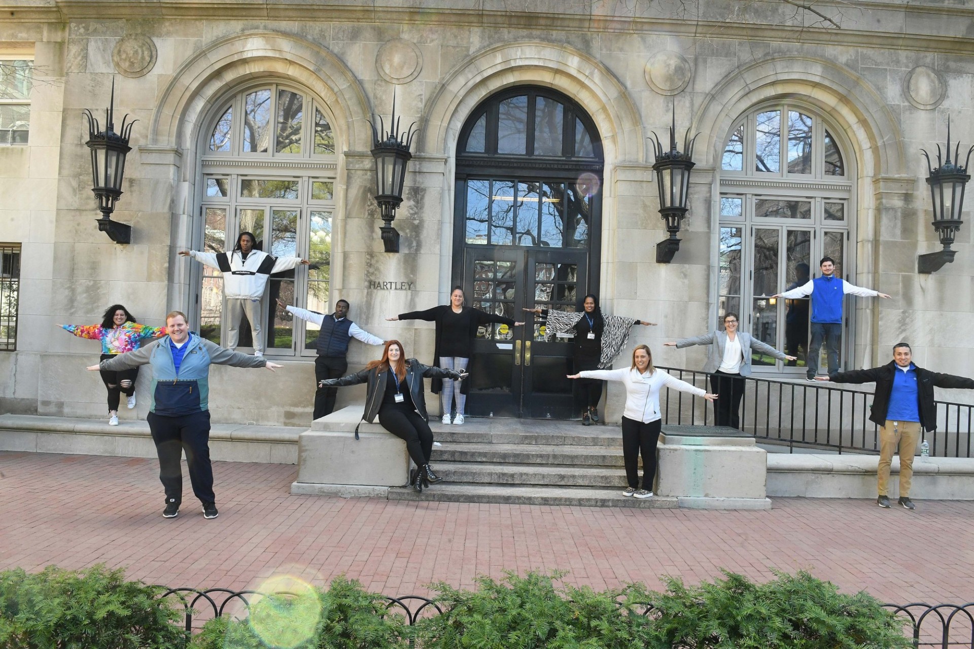 The members of the Housing team demonstrate proper physical distancing technique in front of Hartley Hall. Photo credit Eileen Barroso. Eleven people stand staggered, 6 feet apart from each other, in front of a grey stone building