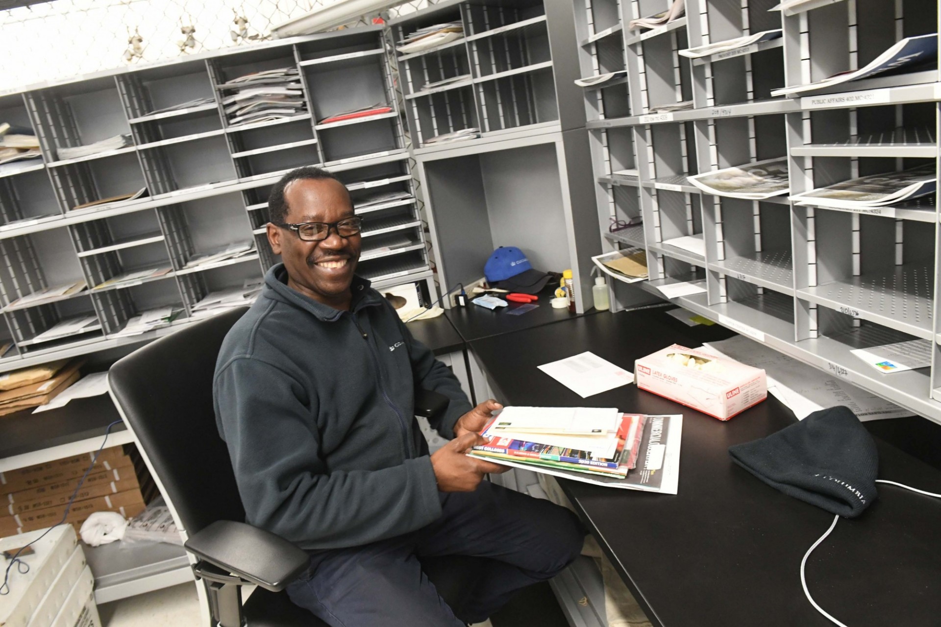 A man with black glasses and a grey jacket sits in a mail room holding a stack of mail.