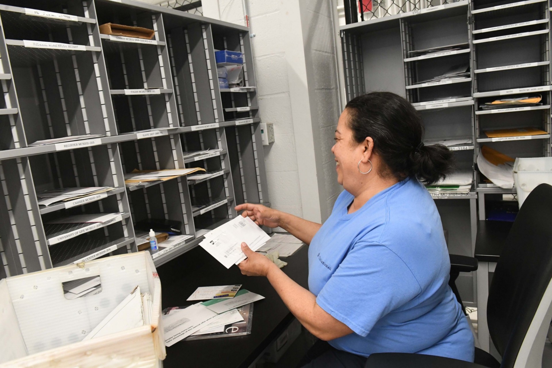 A woman in a blue t-shirt sits in a mail room. She is holding envelopes in her hands.