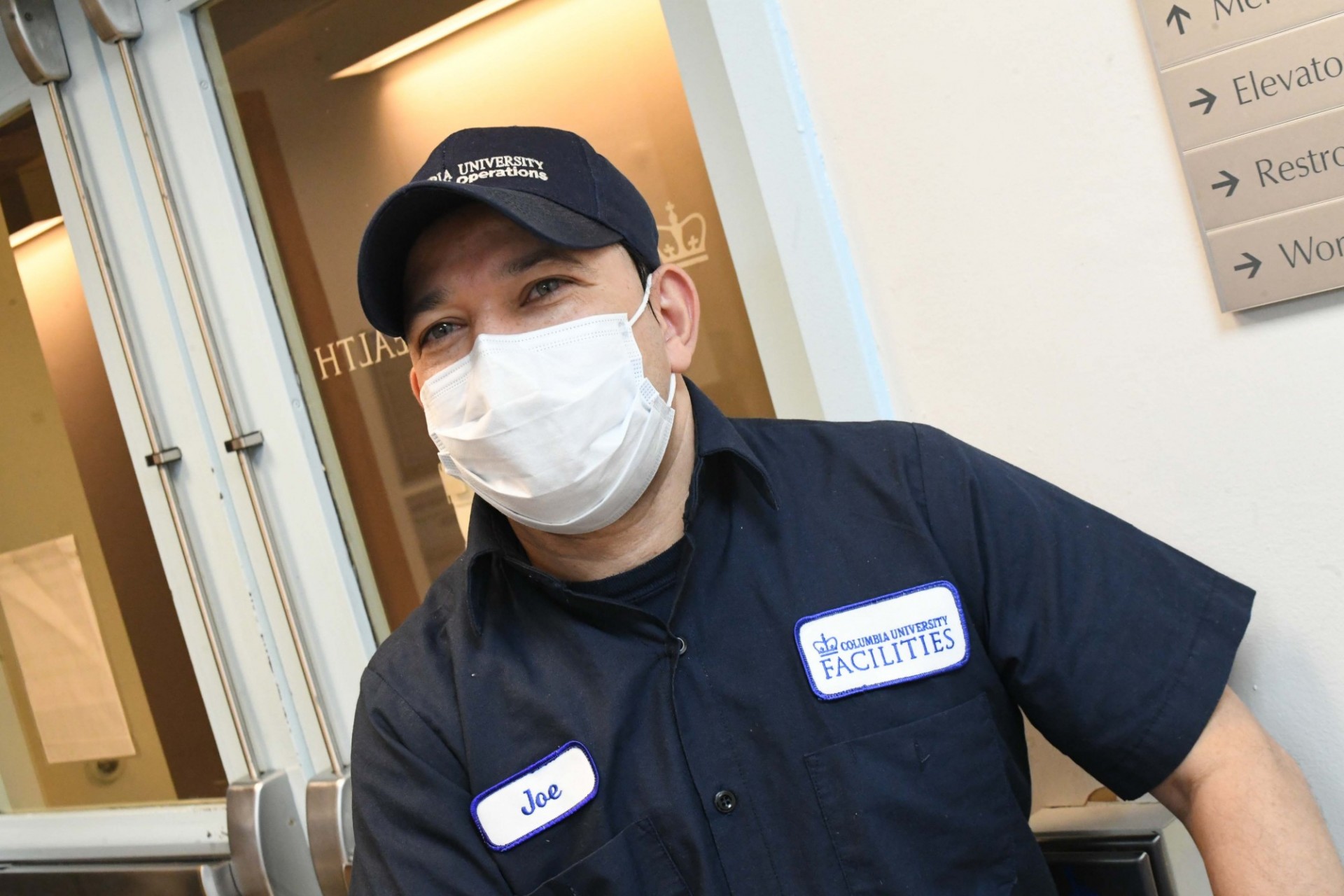 A man in a blue custodial uniform is wearing a mask and standing in front of a doorway.