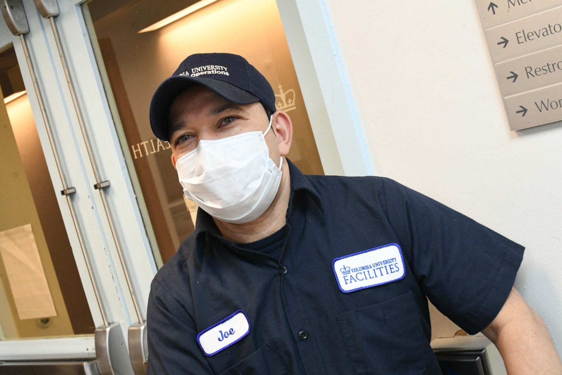 A member of the Custodial team on his rounds. Photo credit Eileen Barroso. A man in a blue custodial uniform is wearing a mask and standing in front of a doorway.