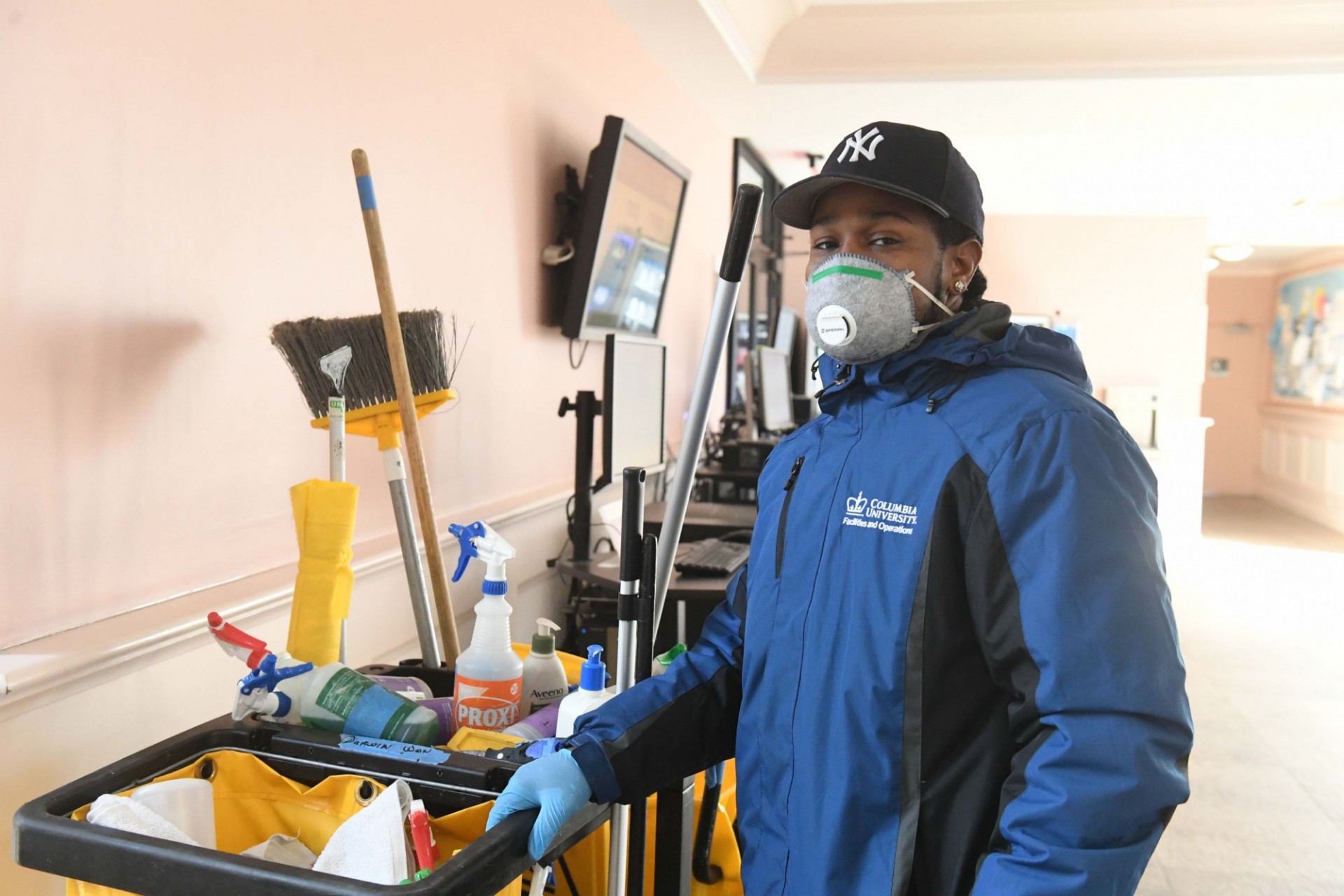 A man wearing a baseball cap and blue jacket wearing a mask stands next to a yellow custodial cart.