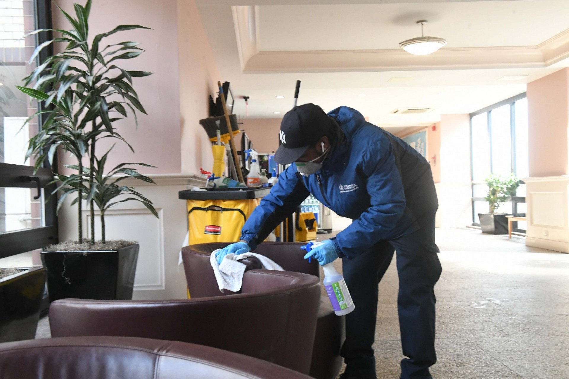 The Custodial team has increased cleanings of high-touch surface areas like communal furniture in building lounges. Photo credit Eileen Barroso. A man wearing a baseball cap and blue jacket wearing a mask and gloves is spraying and wiping down a brown leather chair.
