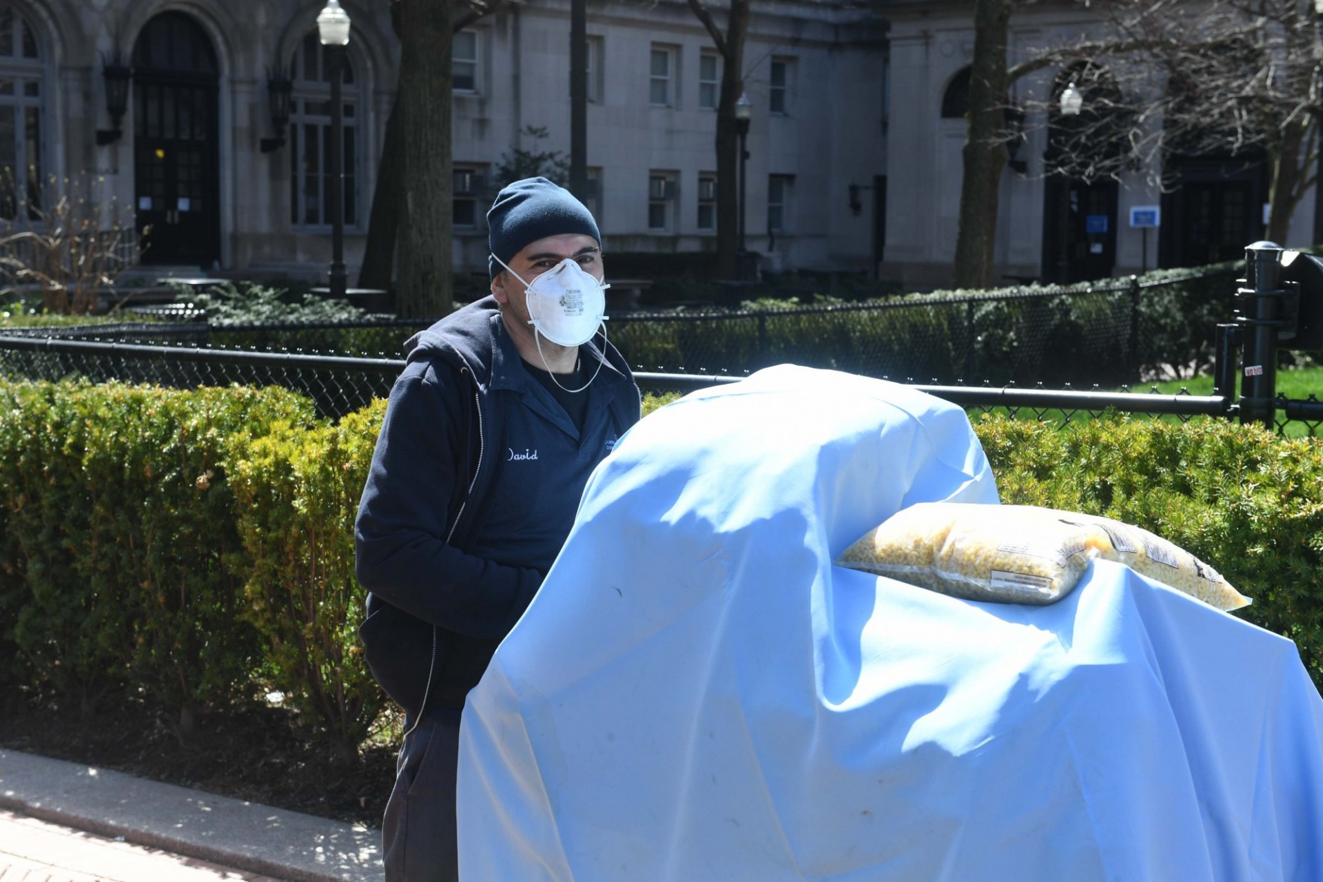 A man in a blue custodial uniform wearing a knit hat, jacket, and mask pushes a covered cart across campus.