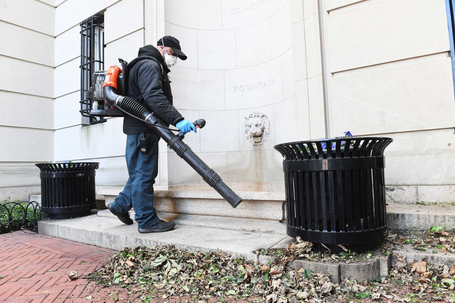A man wearing a mask, baseball cap, and black jacket is equipped with a leaf blower. He is blowing leaves away from a building.