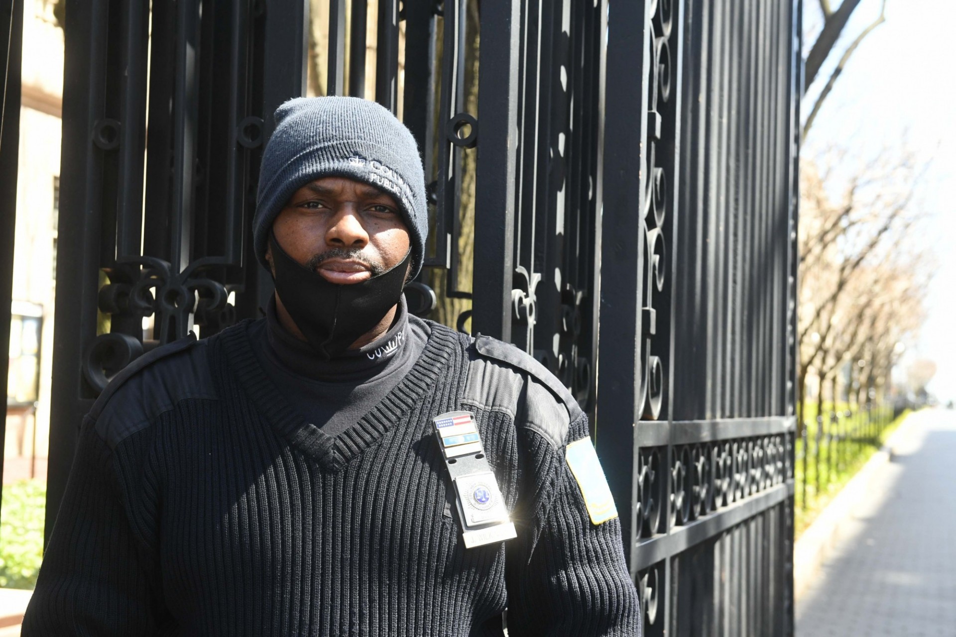 A man wearing a blue beanie and black sweater is standing in front of Columbia's College Walk gates. His black mask is pulled down, revealing his mouth.