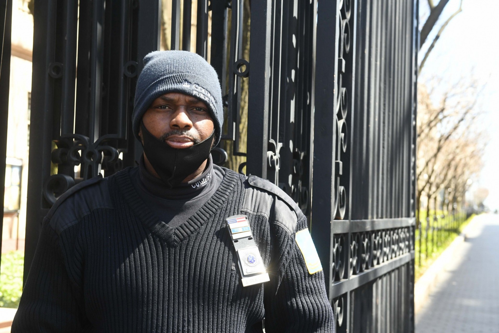 A Public Safety Officer monitors the entrance to College Walk. Photo credit Eileen Barroso. A man wearing a blue beanie and black sweater is standing in front of Columbia's College Walk gates. His black mask is pulled down, revealing his mouth.