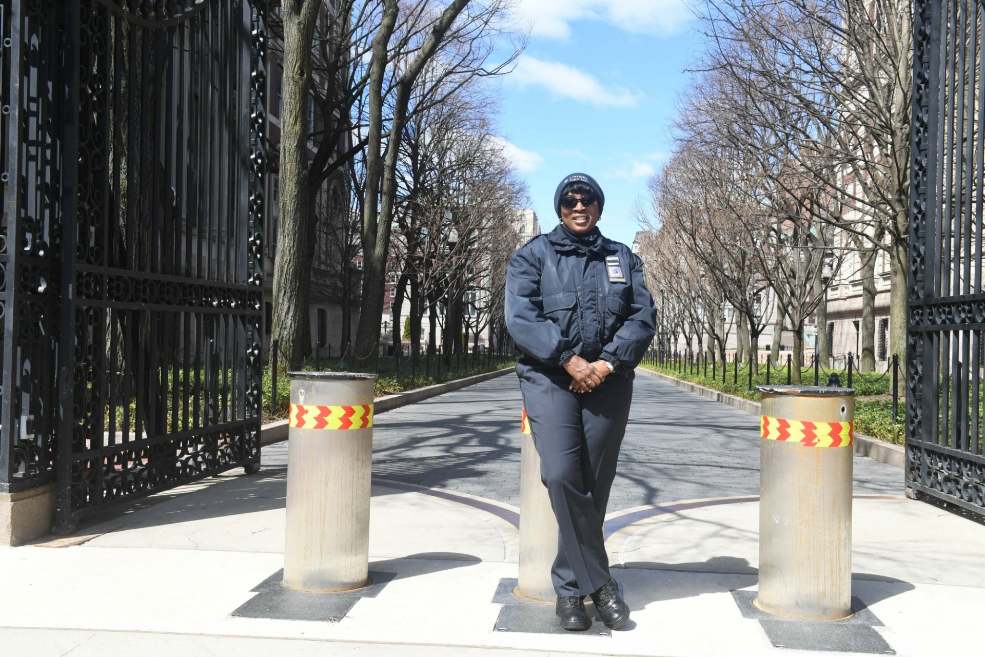 A Public Safety Officer monitors the entrance to College Walk. Photo credit Eileen Barroso. A woman in a blue beanie, sunglasses, and blue jacket leans against a concrete bollard at the entrance of a walkway.