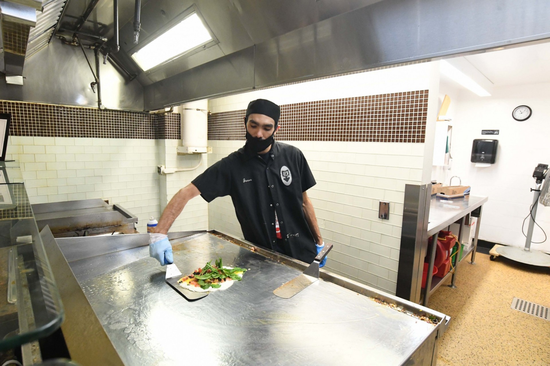 A man in a black Dining uniform wearing a mask prepares food on a griddle.