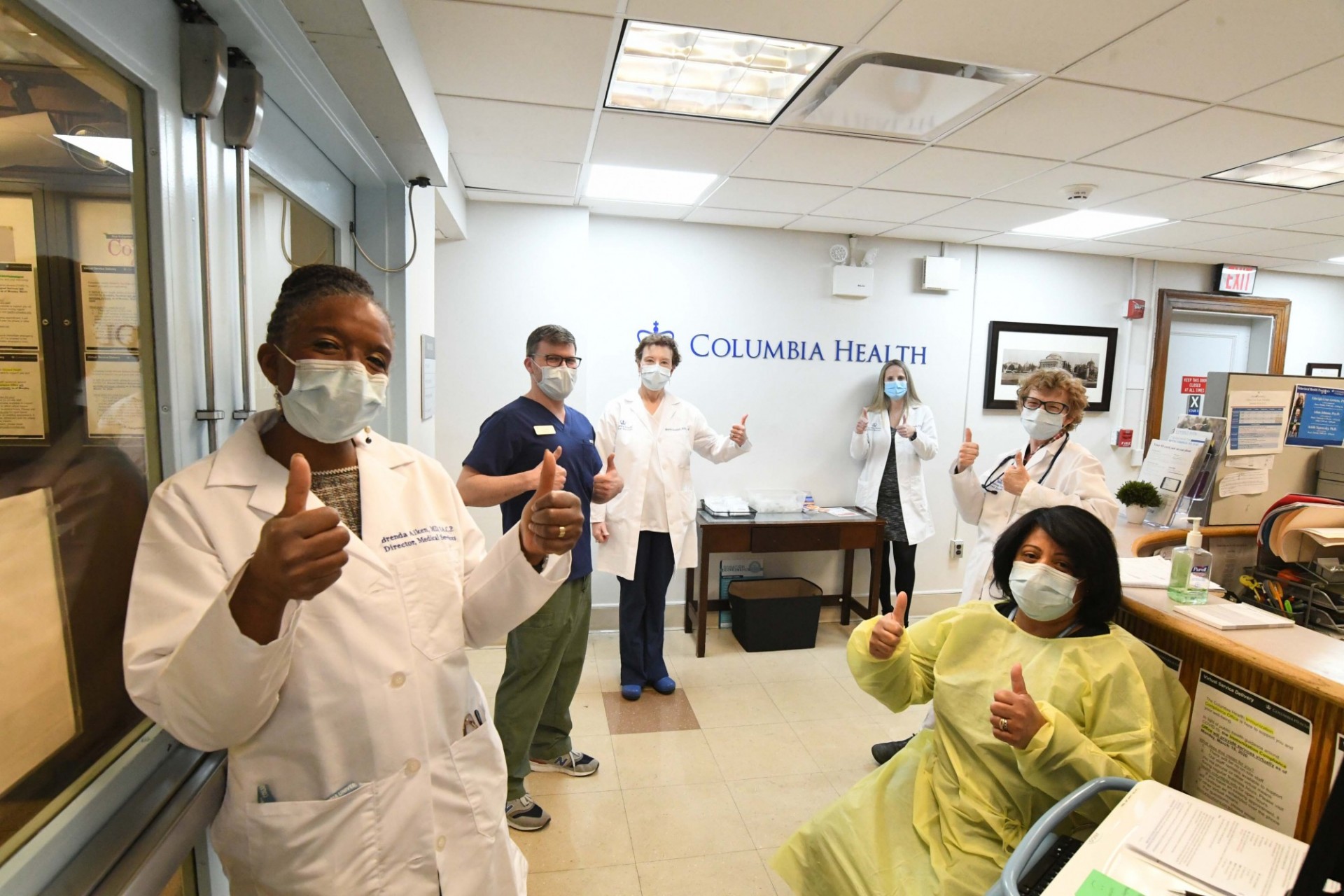 The Medical Services team continues to operate at John Jay serving the non-emergency medical needs of students on campus. Photo credit Eileen Barroso. A group of Columbia Health workers in masks stand in the lobby of the Medical Services unit with thumbs up.