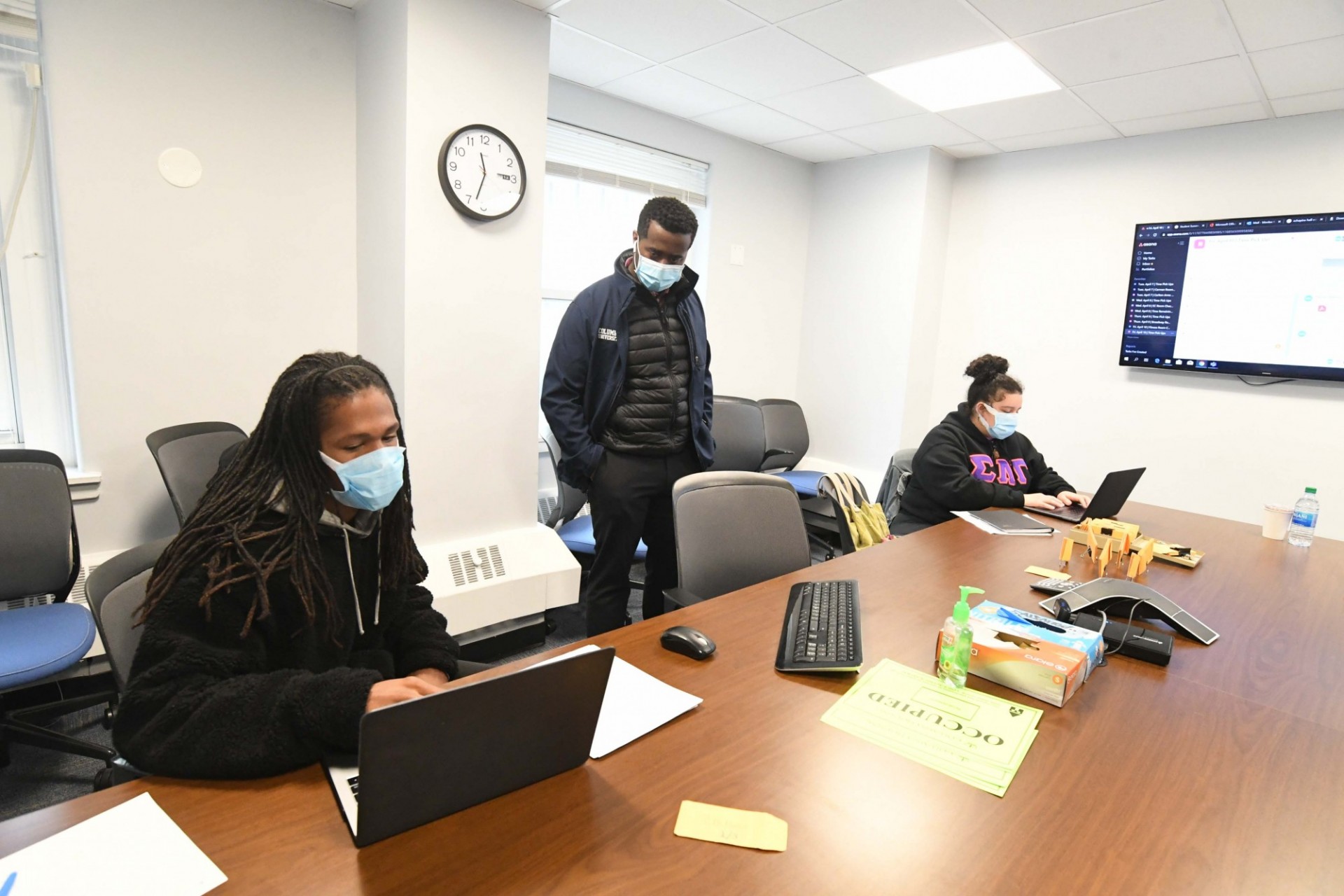 Two people wearing sit at a long conference table working at laptops. In between them, a man wearing a mask is standing up.