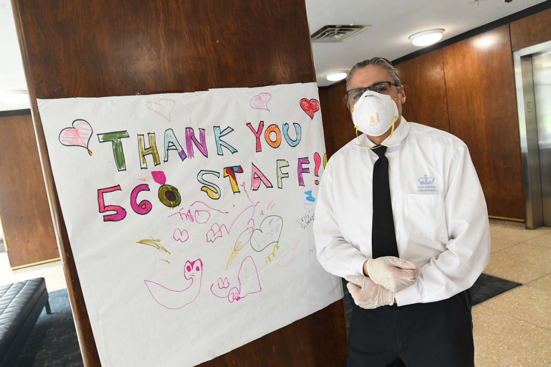 A doorman in a white button-down shirt and black tie stands in the lobby of a residential building next to a homemade sign that says, "Thank you 560 Staff!"