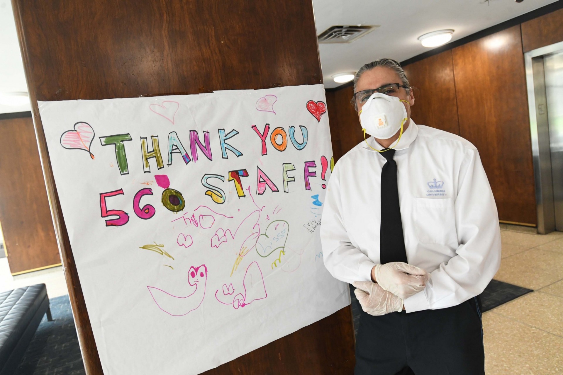The doorman of a Columbia Residential-operated building stands next to a sign made by grateful building tenants. Photo credit Eileen Barroso. A doorman in a white button-down shirt and black tie stands in the lobby of a residential building next to a homemade sign that says, "Thank you 560 Staff!"