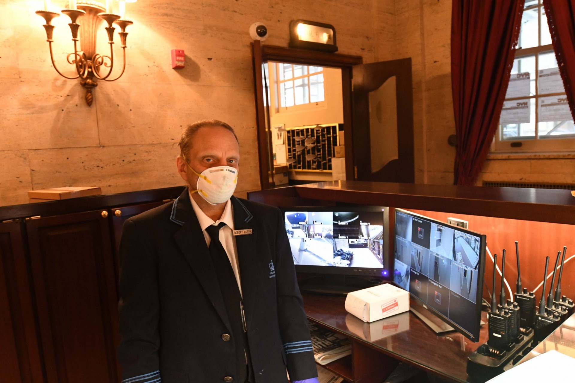 A doorman wearing a black sport jacket stands behind the front desk of a building, next to computer security monitors.