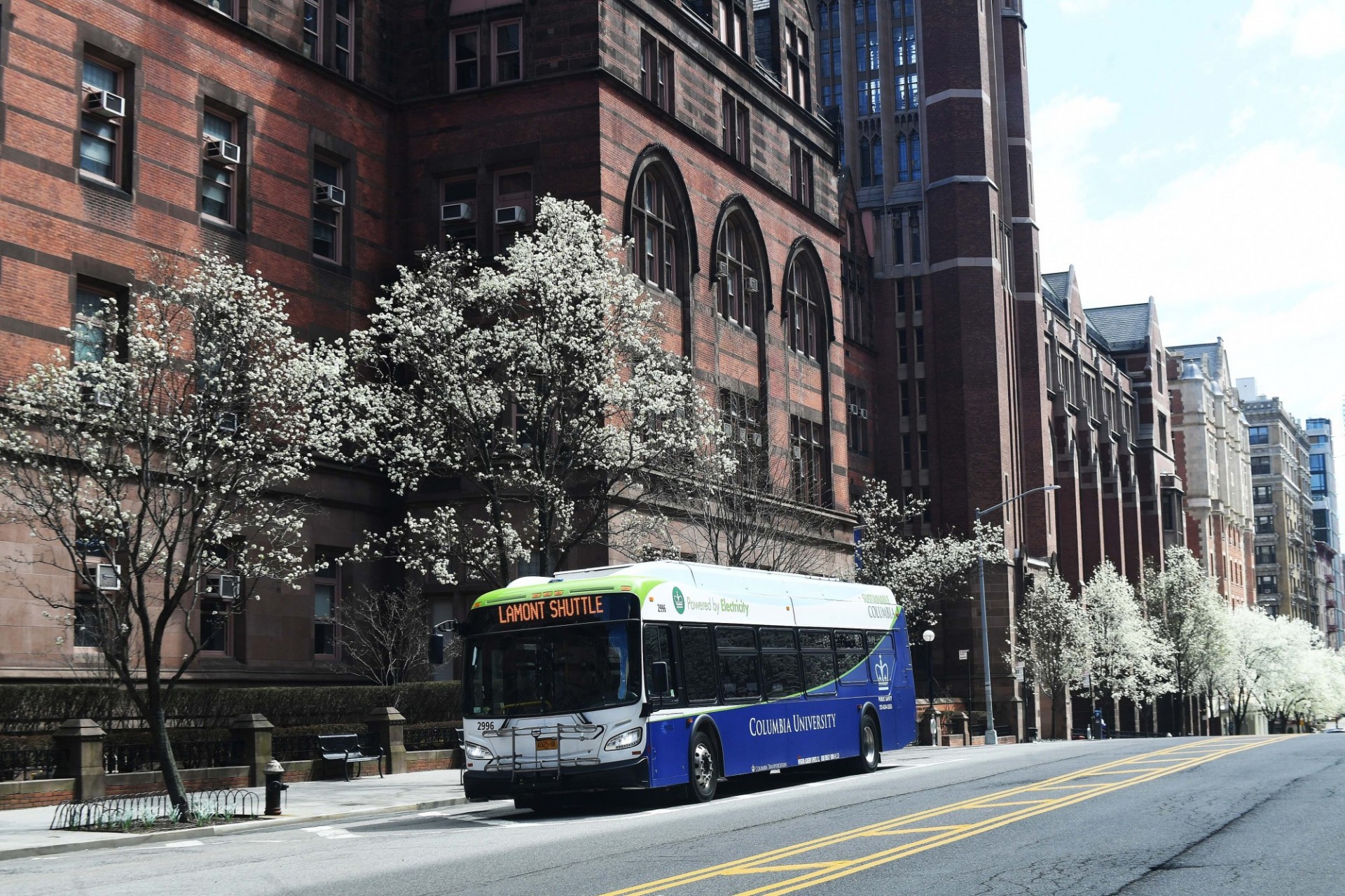 Columbia-operated shuttles continue to provide transportation services. Photo credit Eileen Barroso. A Columbia shuttle bus is pulled over to the side of the road. Behind the bus are trees in bloom and a red brick building.