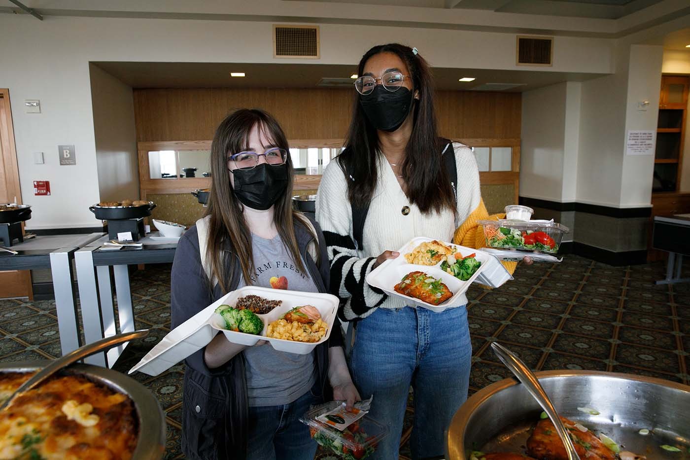 Two students stand behind the serving buffet holding to-go containers.