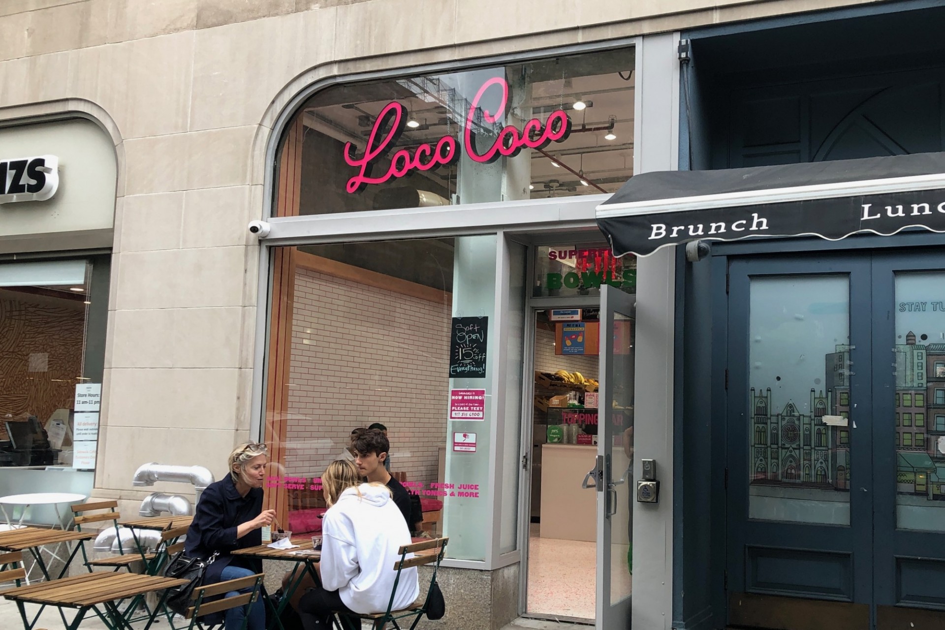 The storefront of Loco Coco, with a sign that says their name in pink cursive font.  A family sits out front and eats their smoothie bowls.