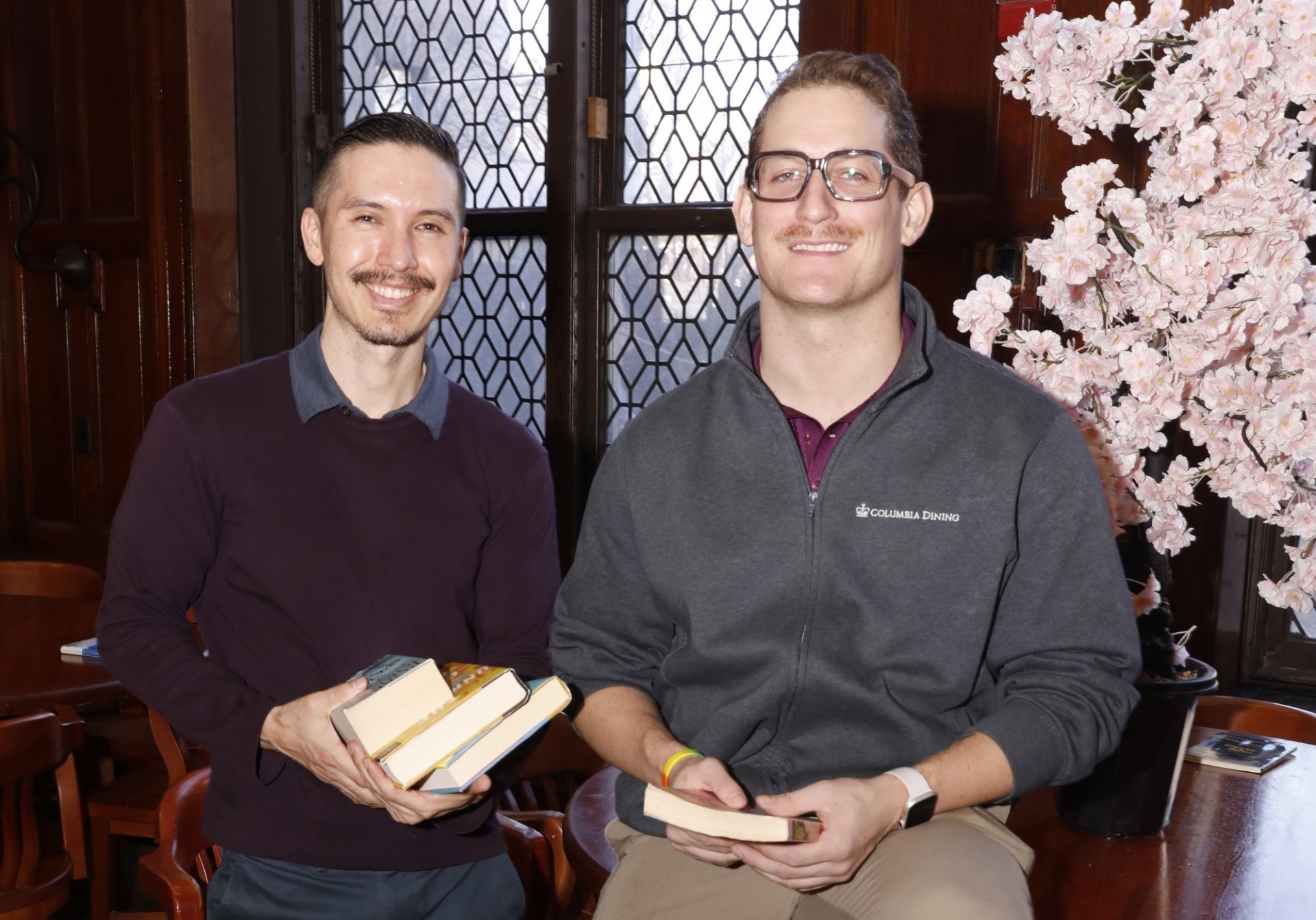 Two people sit indoors by a window; one holds books, the other wears a Columbia Business School sweatshirt and reads. A pink flowering tree is nearby.