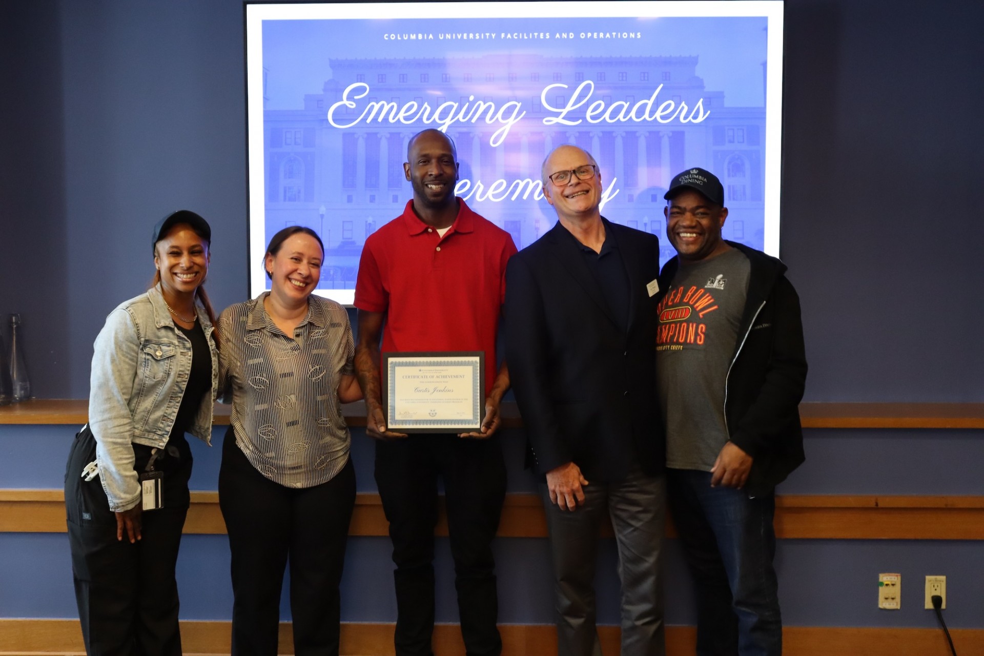 Five people stand in front of a screen reading 'Emerging Leaders Ceremony' at Columbia University; the center person holds a framed certificate.