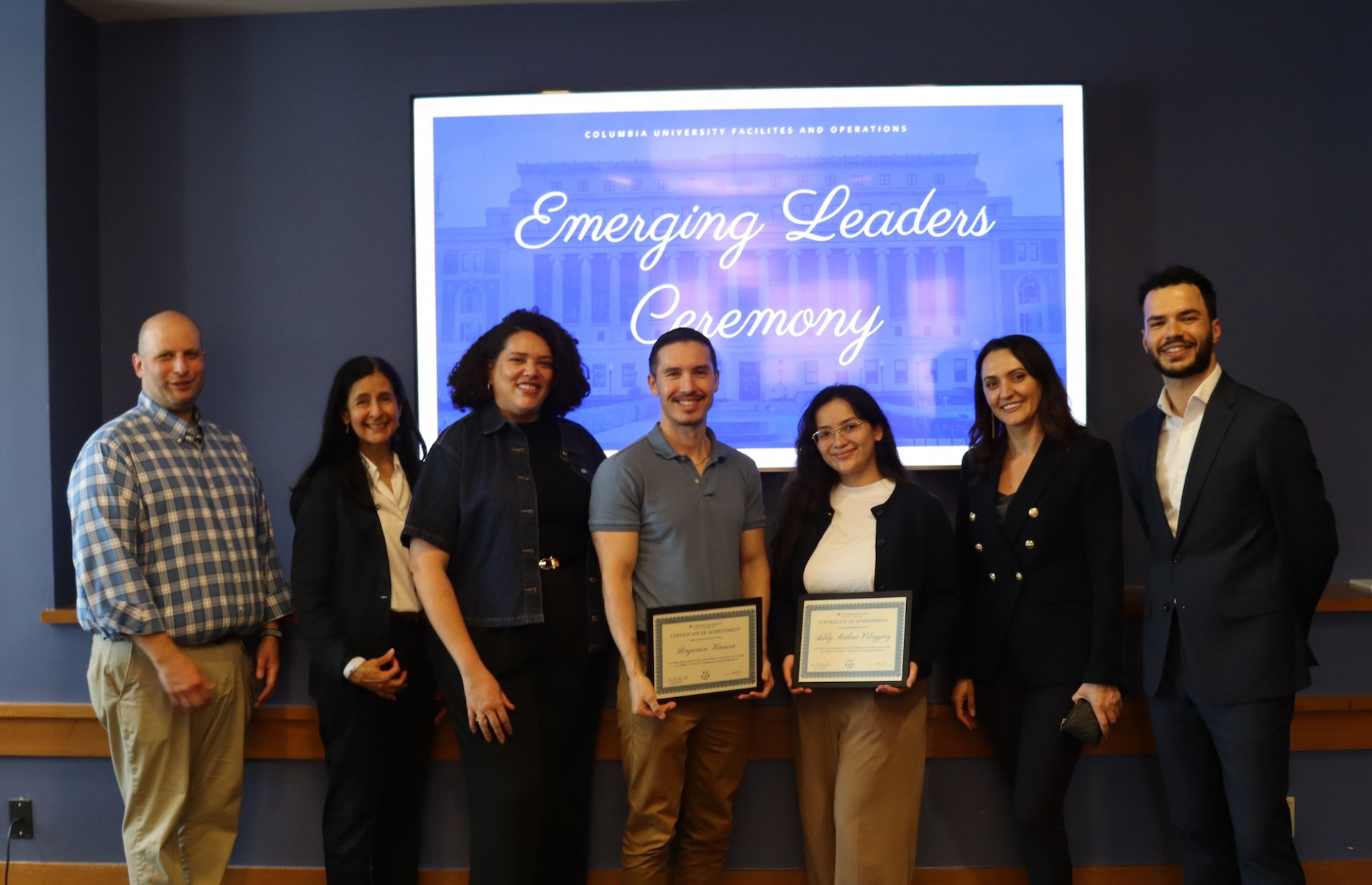 Seven people stand in front of a screen reading 'Emerging Leaders Ceremony' at Columbia University Facilities and Operations; two in the center hold framed certificates.