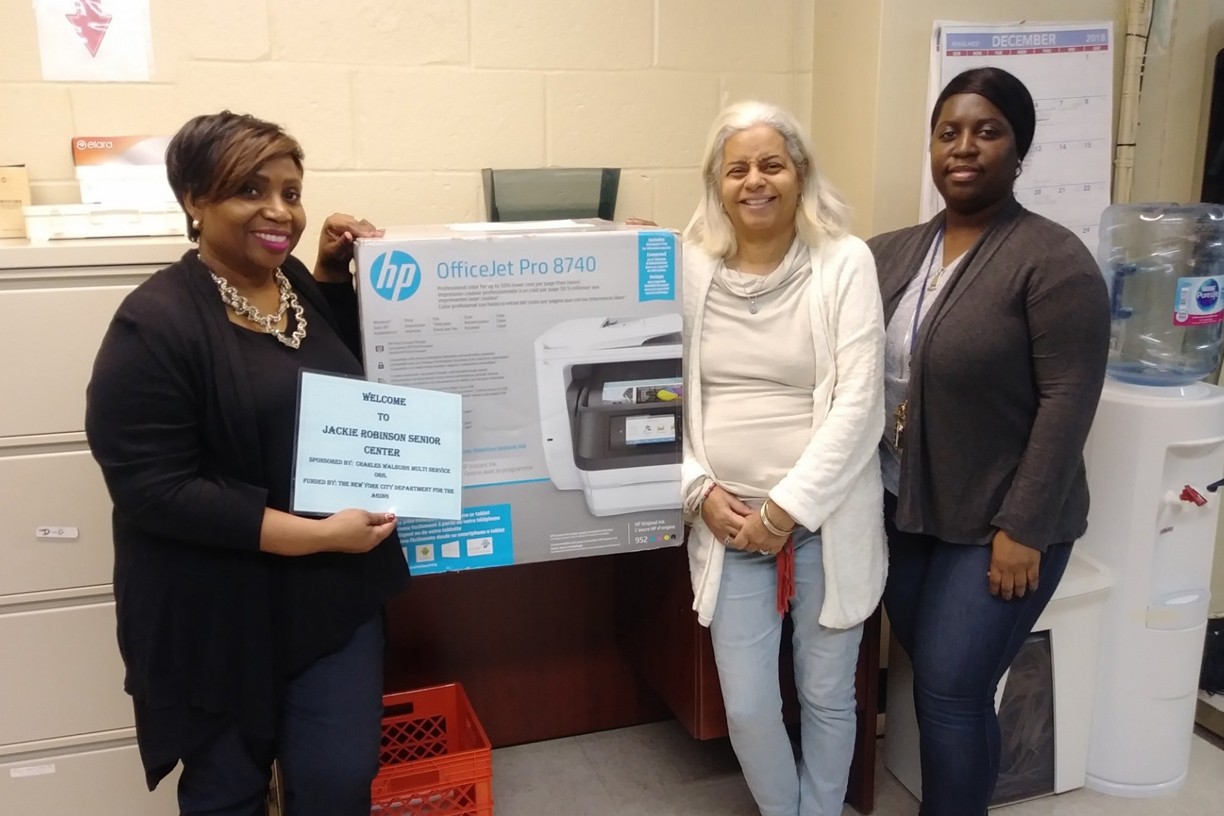 Three women stand next to a brand new printer in its box 
