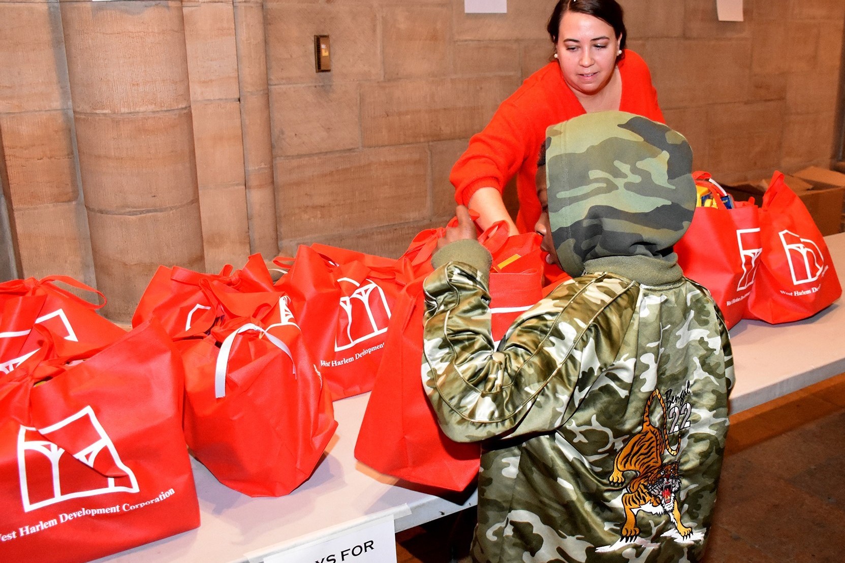 A person hands out a red toy bag to a child standing in front of a table, which is full of toy bags