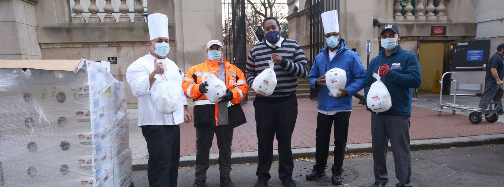 Two chefs and other Columbia Dining team members stand in the street holding turkeys in their hand, next to a pallet full of donated goods