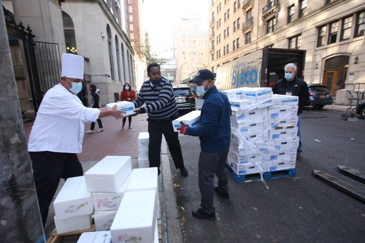 A chef hands a box to two people from a stack of boxes next to him on the street